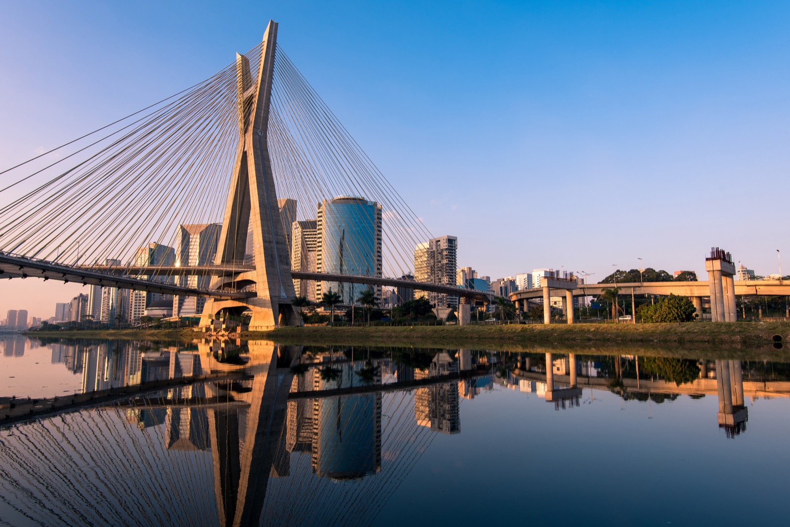Cable-stayed bridge over calm water reflecting modern city skyline, clear sky, and golden sunlight, creating a serene urban scene.